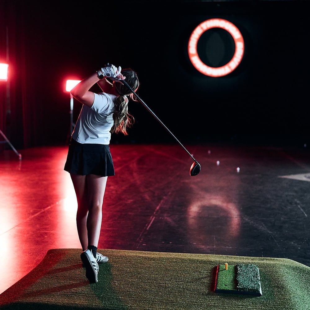 A golfer in a white shirt and black skirt swinging a driver under a red ring light