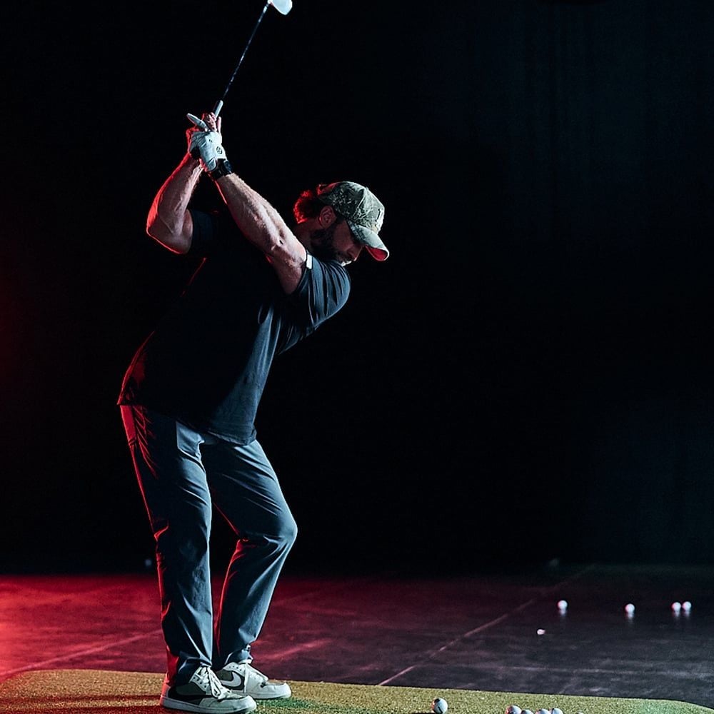 A golfer in a black shirt swinging an iron on the arena turf